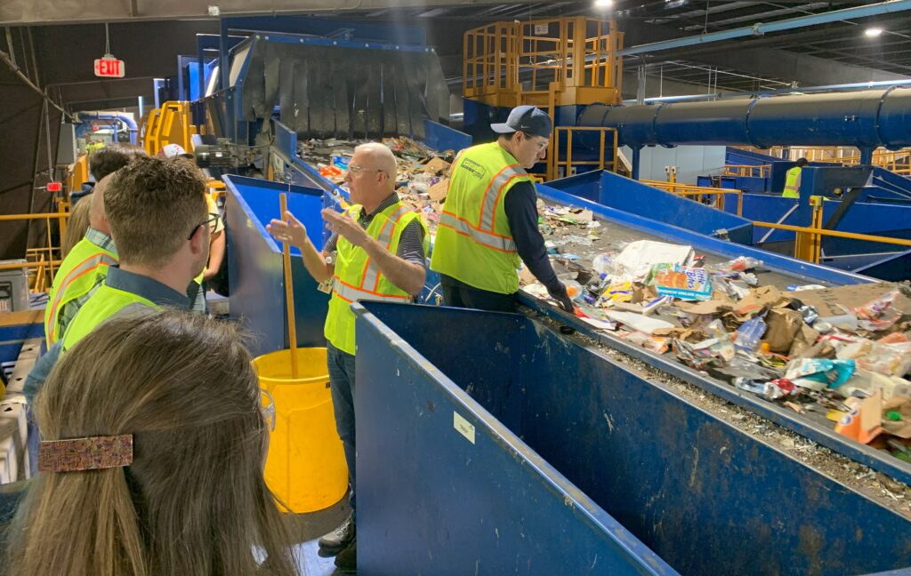 Visitors observe an employee manually sorting recycled materials on a conveyer belt.